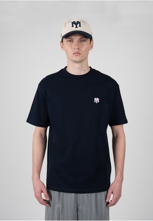 Mladý muž, který má na sobě krémovou a námořnicky modrou čepici a černé tričko s malým logem na levé straně hrudi, doplněné šedými kalhotami s tenkými pruhy.