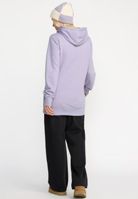 Sweat à capuche violet clair avec poche kangourou, associé à un pantalon noir ample et un bonnet en tricot bicolore. Design décontracté, texture douce.