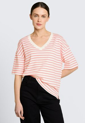 Kobieta ubrana w luźną, białą koszulę z dekoltem w serek, z cienkimi koralowymi poziomymi paskami, oraz czarne spodnie, stojąca na tle jednolitego jasnego tła.