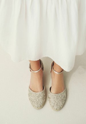 Füße in beigen bestickten Ballerinas mit Knöchelriemen unter einem weißen, fließenden Rock auf einem hellen Boden.