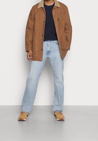 Brun bomulljacka med en beige krage, marinblå tröja under, ljusblå jeans och bruna sneakers med vita accenter. Avslappnad ensemble.