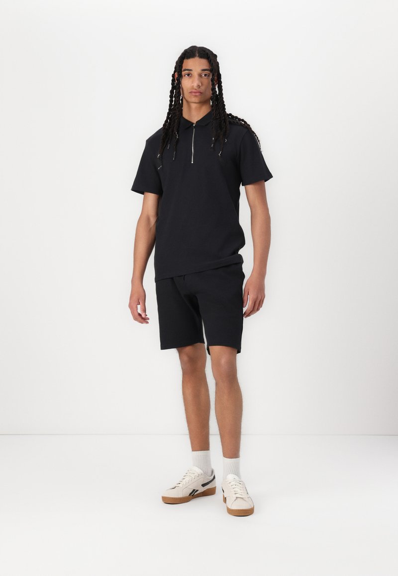 Svart kortermet polo med glidelås krage, kombinert med svarte shorts. Fottøyet har en kremfarge med brun såle og svarte detaljer.