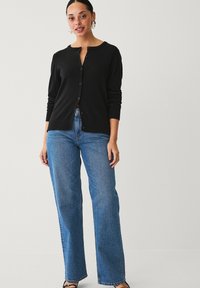 Cardigan nero con scollatura tonda e chiusura a bottoni. Abbinato a jeans a gamba larga blu e sandali neri. Tessuto liscio, vestibilità casual.