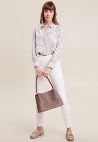 Kastanienbraune Lederhandtasche mit runder Form, geflochtenem Griff und glatter Textur. Das Modell kombiniert sie mit einem gestreiften Shirt und weißen Hosen.