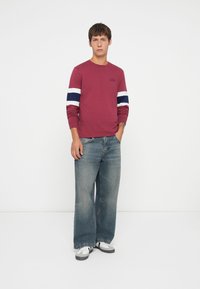 Long-sleeve burgundy shirt with navy and white stripes on sleeves, paired with loose light blue jeans and gray sneakers.