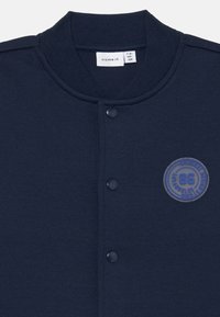 Veste pour enfants bleu marine avec col côtelé, boutons-pression au centre et patch rond indiquant "86 Los Angeles Original Design Studio."