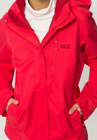 Veste imperméable rouge vif avec fermeture éclair à l'avant, capuche ajustable et deux poches latérales. Présente un logo sur le haut de la poitrine à gauche.