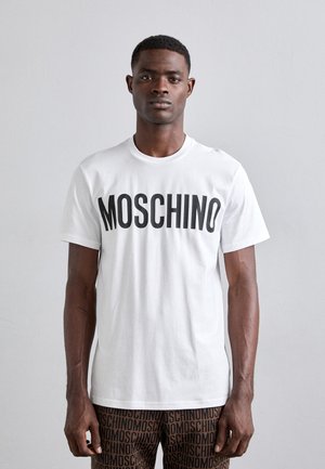 Tricou alb din bumbac cu text masiv negru "MOSCHINO". Modelul poartă pantaloni maro cu un model ce repetă brandingul. Design simplu, cu guler tip crew.