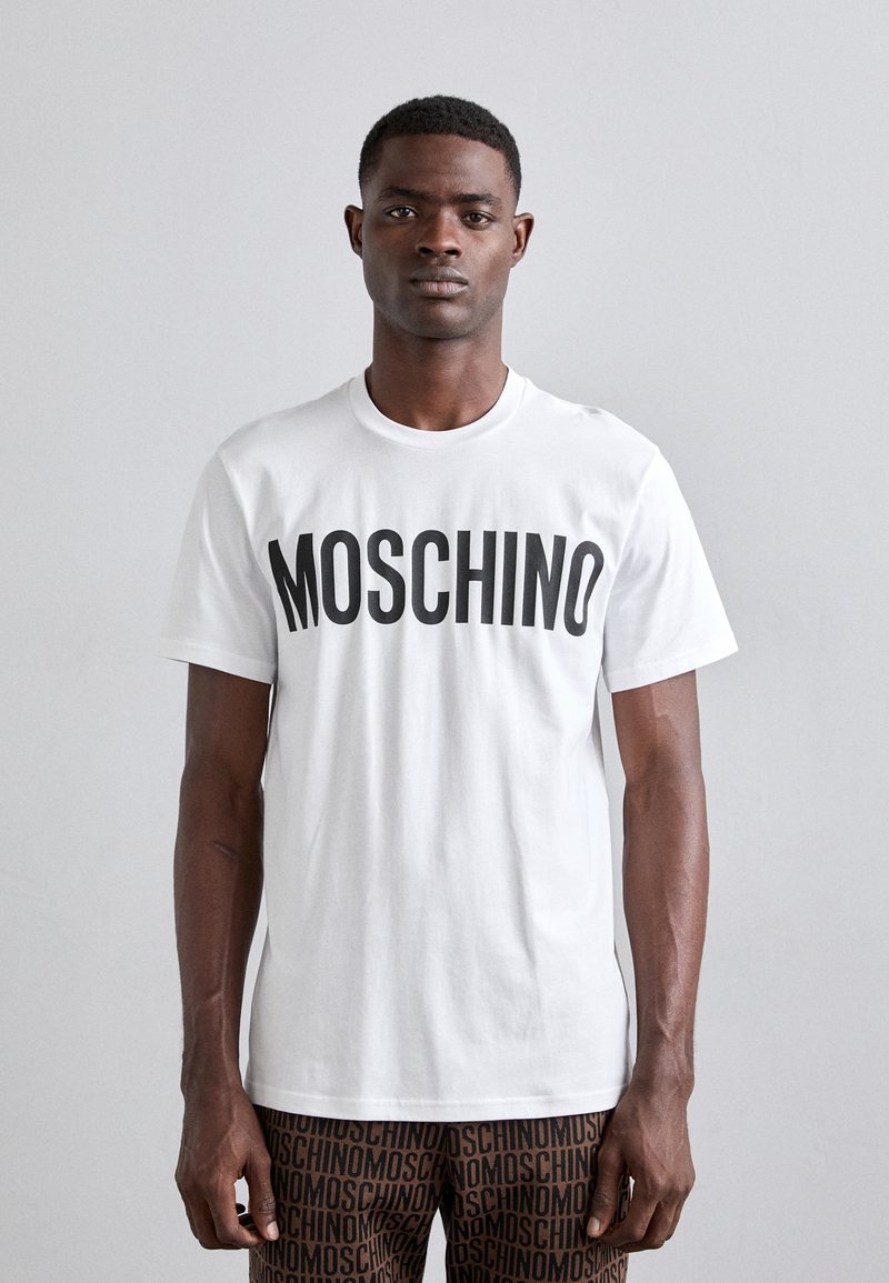Balta medvilninė marškinė su ryškiu juodu "MOSCHINO" tekstu. Modelis vilki raštuotas rudas kelnes su pakartotiniu ženklinimu. Paprastas, suapvalinto kaklo dizainas.
