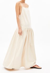 Beige hihaton mekko, jossa on vartalonmyötäinen yläosa ja laskeutuva, kerrostettu hame. Materiaali vaikuttaa pehmeältä; valmistettu minimalistisella designilla ja ohuilla hihnoilla.