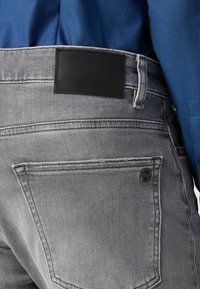Jeans de mezclilla gris con un parche de cuero negro etiquetado "BOSS" en la cintura. El bolsillo trasero presenta un acento negro con un detalle de logo.