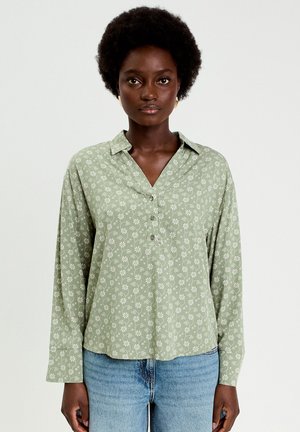 Grünes Hemd mit Blumenmuster aus leichtem Stoff, mit Reverskragen, Knopfleiste vorne und langen Ärmeln; kombiniert mit blauen Jeans.