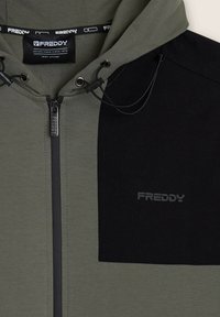 Sudadera con capucha de color verde oliva oscuro y negro, cierre con cremallera, capucha con cordón, adorno de tela y logotipo bordado en el pecho.