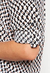 Svart- och vitt mönstrad skjorta med cirkulära mönster och persikofärgade accenter. Tyget är mjukt, och manschetten har en enda svart knappdetalj.