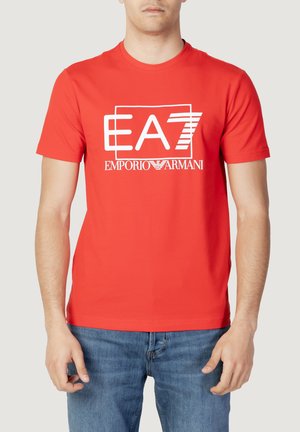 Mężczyzna noszący jaskrawoczerwoną koszulkę z krótkim rękawem z białym logo "EA7 Emporio Armani", w połączeniu z niebieskimi dżinsami, pokazany od szyi do bioder.