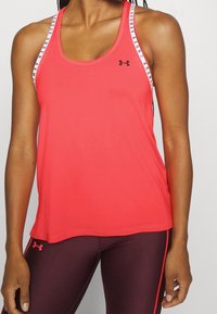 Vrouw draagt een mouwloze rode Under Armour tanktop en bordeauxkleurige leggings met rode zijstrepen, staande tegen een effen achtergrond.