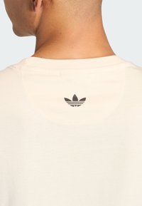 Svijetlo bež majica s okruglim izrezom i crnim Adidas logom na gornjem dijelu leđa. Mekana tkanina s glatkom teksturom.