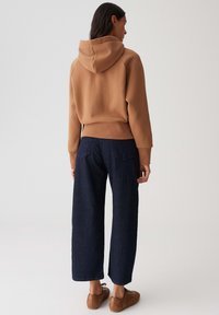 Vrouw in een chocoladebruine hoodie, met een donkere brede spijkerbroek en bruine sneakers, staat met haar rug naar de camera tegen een effen achtergrond.