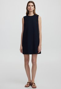Vestido sin mangas, hasta la rodilla, en tejido de un oscuro color azul marino con una textura suave. Presenta un diseño minimalista y un detalle decorativo de botón en un hombro.