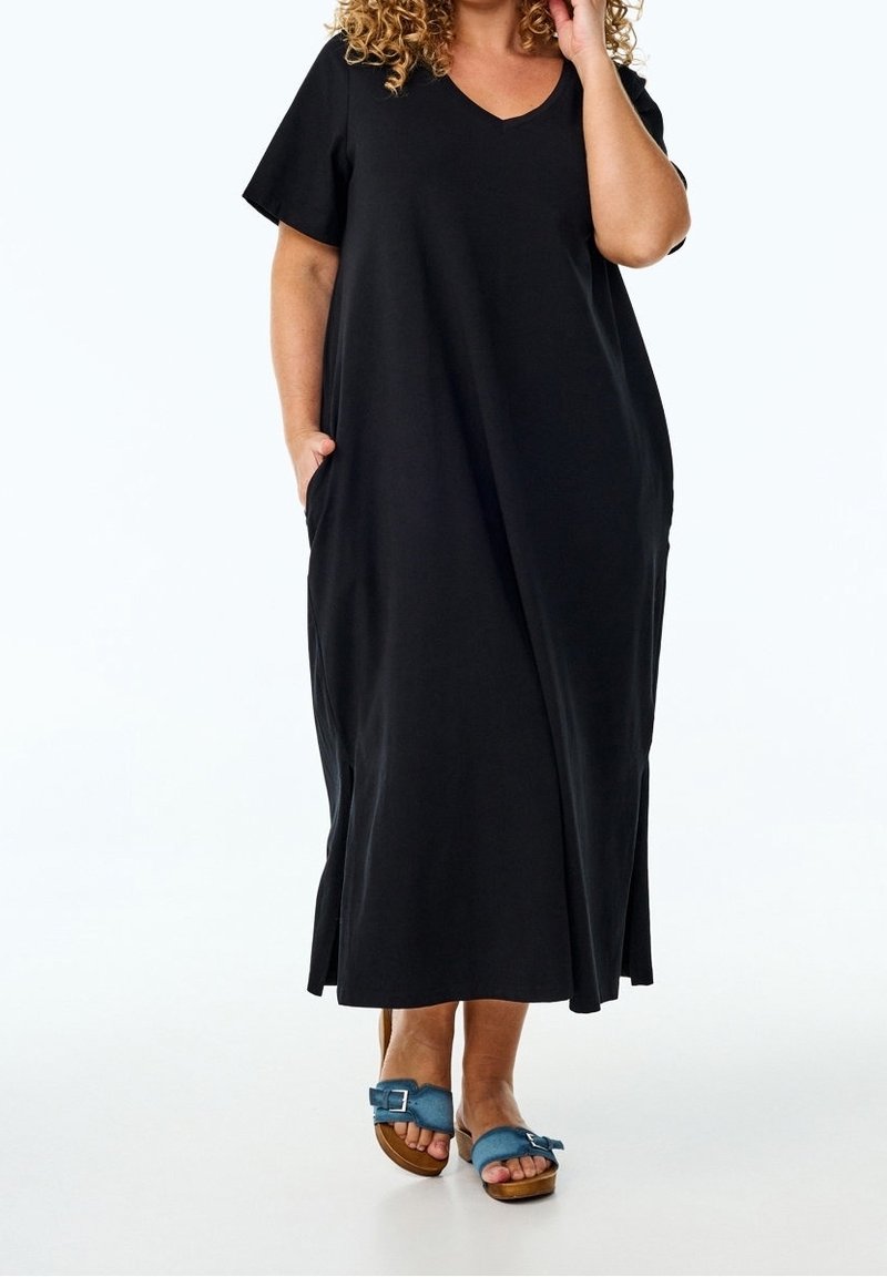 Femme portant une robe maxi noire à manches courtes et des sandales ouvertes bleues, debout avec une main dans la poche de la robe, fond blanc.