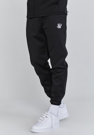 Person iført sorte tætsiddende joggers med hvidt logo på låret og hvide sneakers med tykke snørebånd på en lys baggrund.