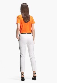 Pomarańczowa bawełniana koszulka i białe dopasowane spodnie z czarnymi bocznymi paskami. Modelka ma na sobie czarne sandały na słupku. Widok z tyłu.