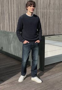 Jeune homme debout à l'extérieur, les mains dans les poches, portant un pull foncé, un jean bleu et des baskets blanches devant un mur en bois.