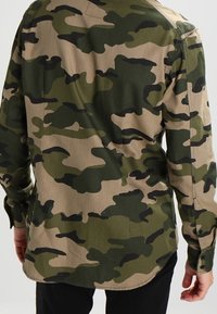 Camo-shirt met een mix van groene, zwarte en beige patronen, gemaakt van lichtgewicht stof met lange mouwen en knoopmanchetten.