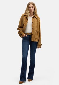 Bruine suède jas met grote zak en knoopsluiting, gecombineerd met nauwsluitende donkerblauwe uitlopende jeans en tan hakken. Model staat tegen een witte achtergrond.