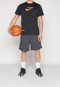 Černé tričko Nike s oranžovým logem, šedé šortky s akcenty NBA a Nike, černobílé sportovní boty, držící oranžový basketbal.