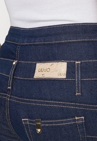 Jeans de mezclilla azul oscuro con cintura alta, costuras doradas y una etiqueta de cuero de marca. Incluye un pequeño acento metálico en el bolsillo trasero.