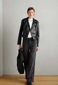 Persoon in een zwart leren jack, witte top, donkere wijde jeans, zwarte schoenen, met een zwarte tas, die binnenshuis loopt op een houten vloer.