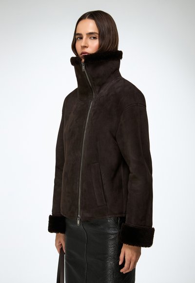 Brązowa zamszowa kurtka z wysokim kołnierzem, zamkiem błyskawicznym i pluszowymi futrzanymi mankietami. W połączeniu z czarną teksturowaną spódnicą o eleganckiej sylwetce.