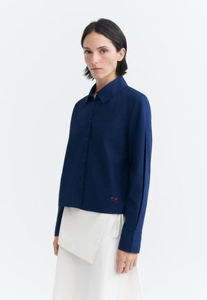 Femme aux cheveux courts et foncés portant une chemise bleu marine à manches longues boutonnée et une jupe asymétrique blanche sur un fond blanc uni.