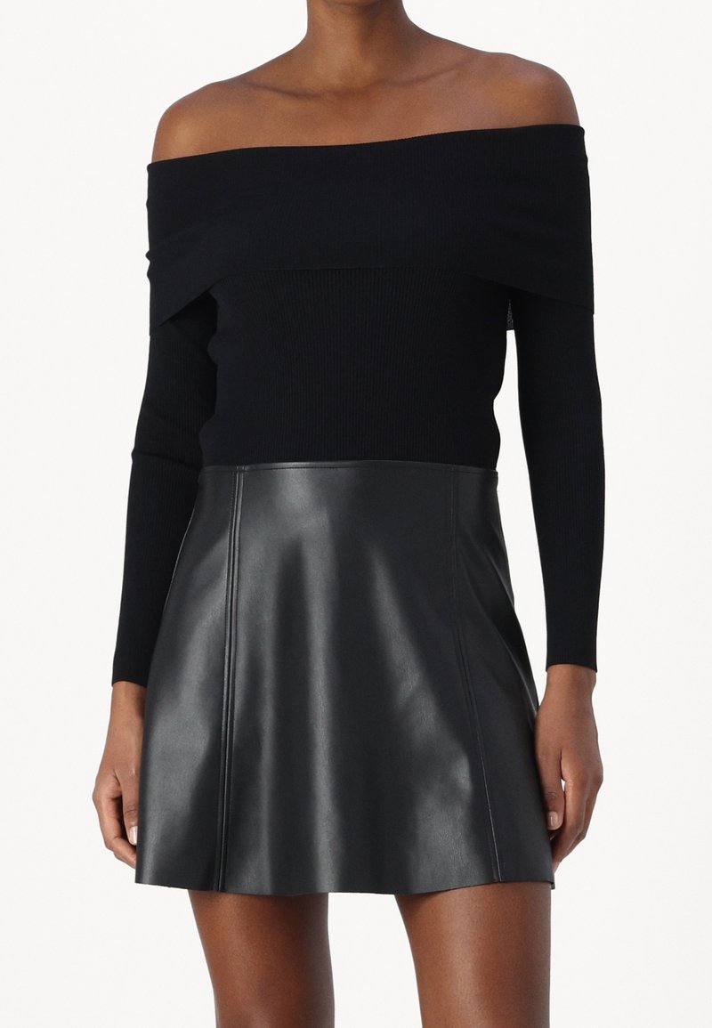 Femme portant un haut noir à manches longues épaules dénudées et une mini-jupe en cuir noire en ligne A, montrée de la mi-cuisse aux épaules.