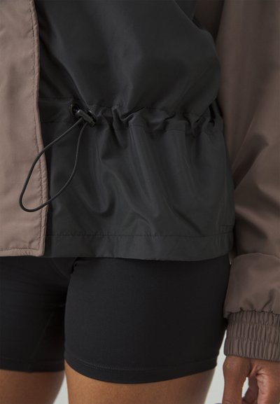 Farbig unterteilte Windjacke mit schwarzen und braunen Abschnitten, mit gerafftem Bund und verstellbarem Kordelzug. Glattes, leichtes Material.