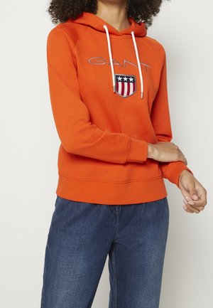 Personne portant un sweat à capuche orange avec des cordons blancs, un logo en forme de bouclier, et un jean bleu, debout avec un bras croisé sur l'autre.