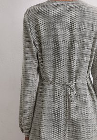 Rückansicht einer Person, die ein gemustertes Kleid mit langen Ärmeln und einem dünnen, gebundenen Gürtel in der Taille vor einer schlichten Wand trägt.