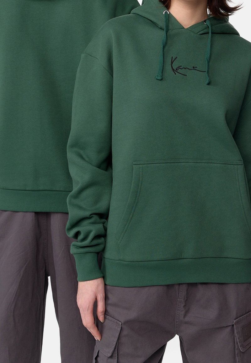 Dos personas vestidas con sudaderas verde oscuro y pantalones cargo gris, con la mano de una de ellas visible descansando a su lado.