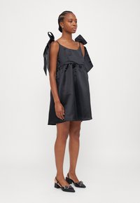 Svart satin klänning med fyrkantig halsringning, rynkad midja och stora rosettdekorationer på axlarna. Parat med svarta slingback-klackar.