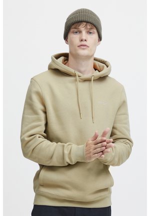Light brown hoodie with a soft texture, drawstring hood, and ribbed cuffs. Paired with a dark knit beanie. Minimal branding on chest.