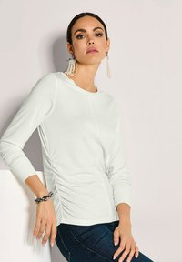 Eine Frau mit dunklen Haaren trägt ein weißes Langarmoberteil, dunkle Jeans, Perlenquastenohrringe und ein schwarzes Perlenarmband und posiert vor einer hellen Wand.