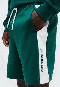 Σκούρο πράσινο σορτς με λευκή πλευρική λωρίδα και το κείμενο "J.LINDEBERG", φορεμένο με το χέρι εν μέρει στην τσέπη και λευκό κορδόνι στη μέση.