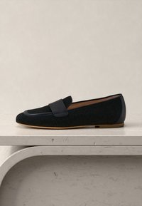 Μαύρο loafer από δερμάτινη υφή με επίπεδη καφέ σόλα, τοποθετημένο σε επιφάνεια από ανοιχτόχρωμο μάρμαρο με ουδέτερο φόντο.