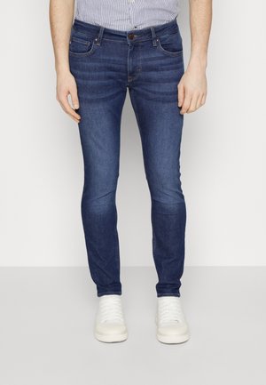 Homme portant un jean slim bleu foncé, des baskets blanches et une chemise rayée rentrée, debout devant un fond uni.