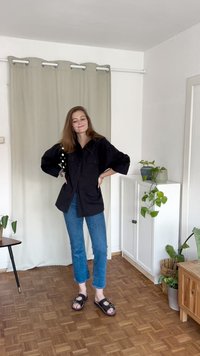 Lockere schwarze Hemdbluse mit zwei Taschen, getragen über blauen Jeans; schwarze Sandalen mit Schnalle; Innenraum mit Pflanzen und Holzboden.