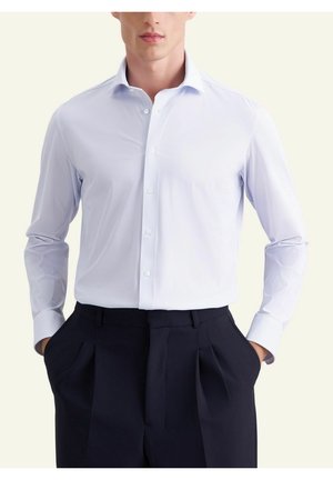 Homme portant une chemise boutonnée texture bleu clair rentrée dans un pantalon plissé taille haute bleu marine foncé, les mains dans les poches.