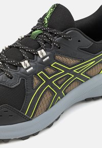 Zwarte sportschoen van mesh en synthetische materialen, met een grijze zool, groene en gele accenten, en een gestructureerd ontwerp voor duurzaamheid.
