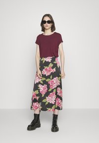 Top curto bordeaux de mangas curtas combinada com uma saia maxi preta com padrão floral, apresentando flores rosas e folhas verdes; botas de combate pretas com atacadores.