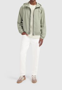 Lichtgroene nylon jas met een capuchon, elastische manchetten en een ritssluiting aan de voorkant, gecombineerd met een crèmekleurig shirt en witte broeken. Bruine sneakers.